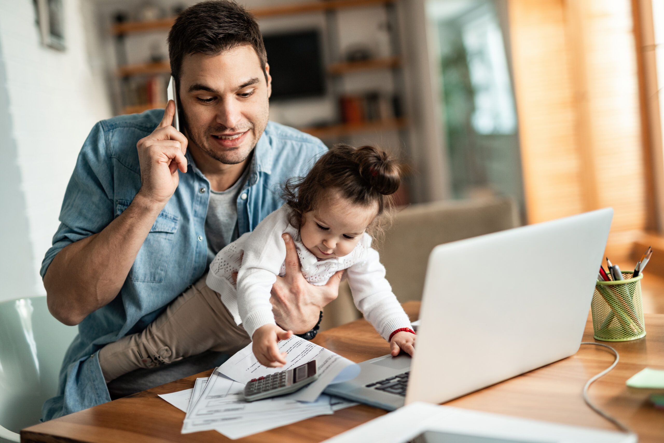 Image of a parent holding child, whilst on the phone and using a laptop