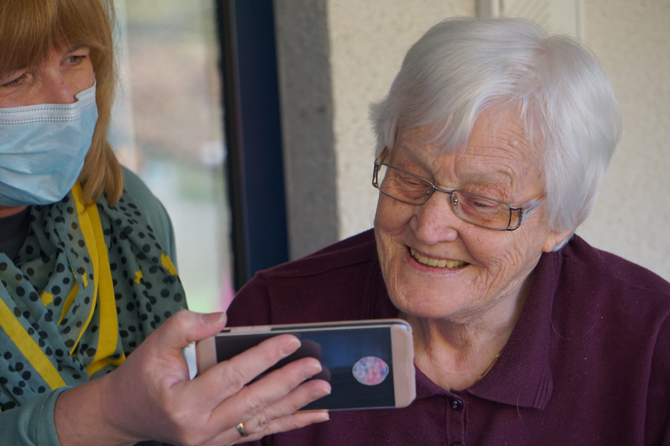 An image of an older lady smiling whilst looking at something on another lady's phone